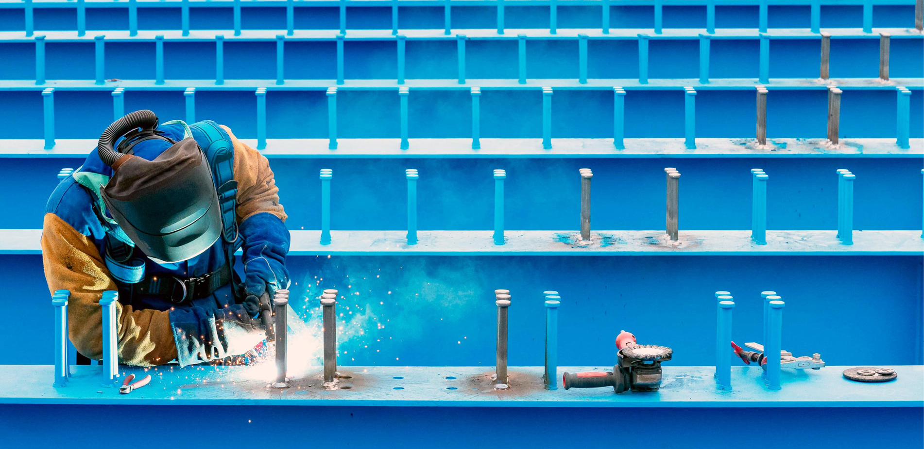Eine Person tr gt Schutzkleidung und einen Schwei helm und schwei t an einer blauen Metallkonstruktion in einem Bereich mit blauen Sitzreihen im Hintergrund. Beschreibung von KI generiert