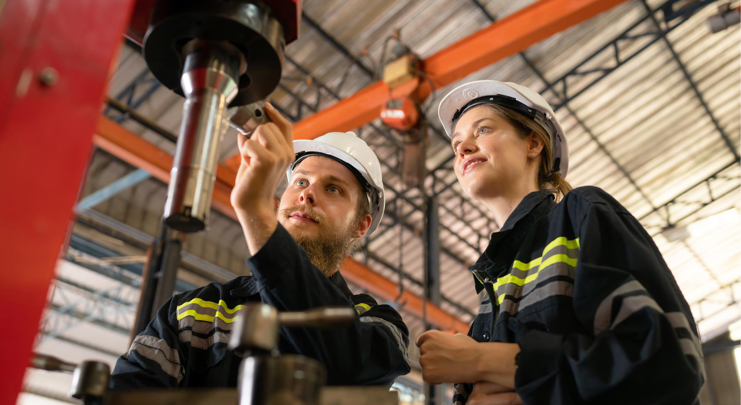 Zwei Personen in Schutzhelmen und Arbeitskleidung bedienen gemeinsam eine Maschine in einer Industriehalle. Beschreibung von KI generiert