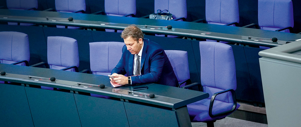 Eine Person in einem blauen Anzug sitzt allein in einem gro en, modernen Sitzungssaal mit blauen St hlen und blickt auf ein mobiles Ger t in den H nden. Beschreibung von KI generiert