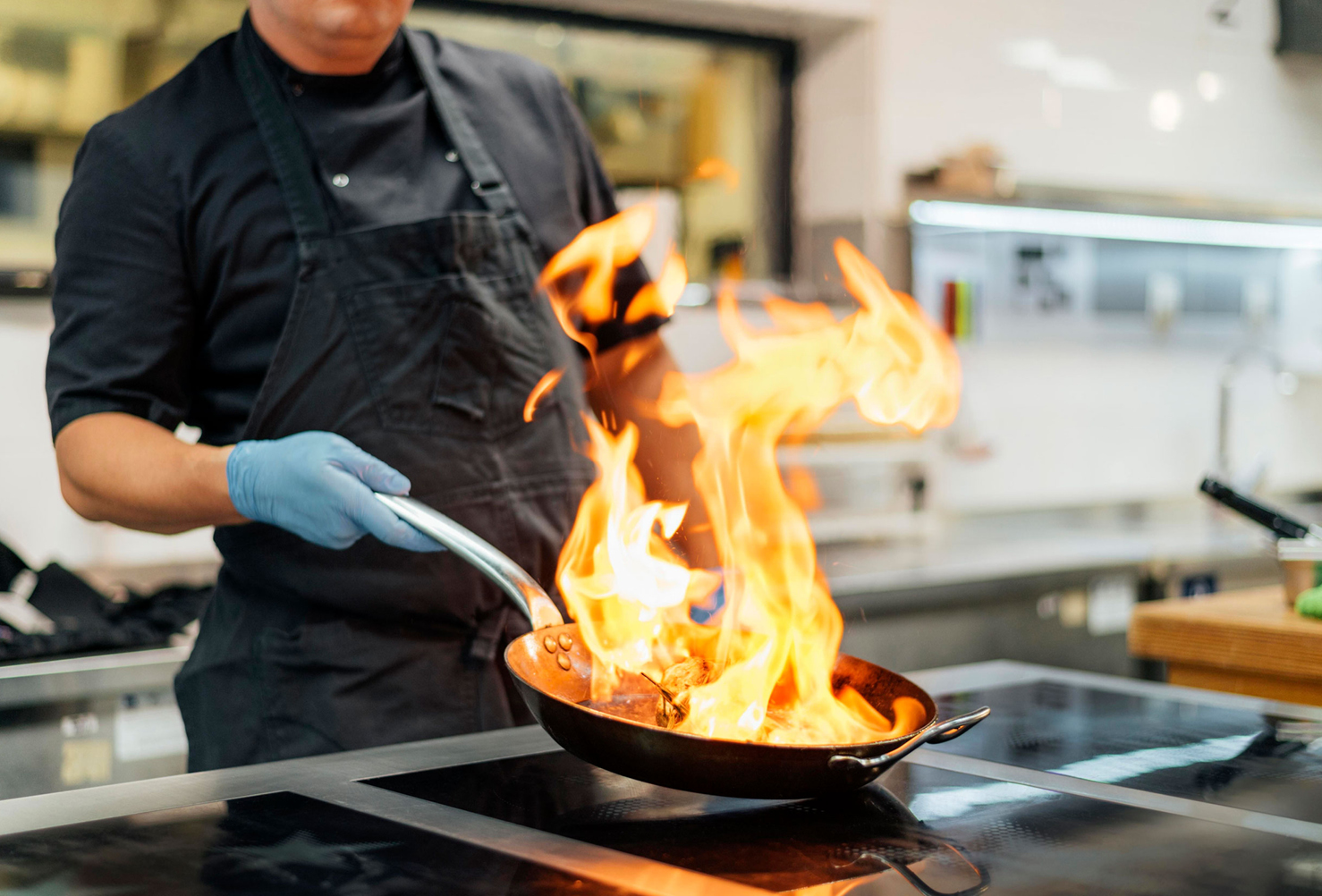 Eine Person in schwarzer Kochkleidung und blauen Handschuhen h lt eine Pfanne mit einer gro en Flamme ber einem Herd in einer modernen K che.  Beschreibung von KI generiert