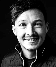 Eine l chelnde Person mit kurzem, zerzaustem Haar tr gt eine Jacke mit offenem Rei verschluss vor einem dunklen Hintergrund. Beschreibung von KI generiert