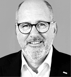 Eine l chelnde Person mit Brille, Bart und Anzug vor einem einfarbigen Hintergrund. Beschreibung von KI generiert