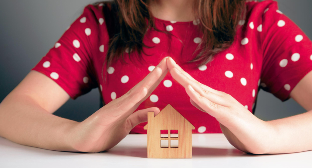 Eine Person in einem roten, gepunkteten Oberteil formt mit den H nden ein Dach ber einem kleinen Holzhaus auf einem wei en Tisch.  Beschreibung von KI generiert