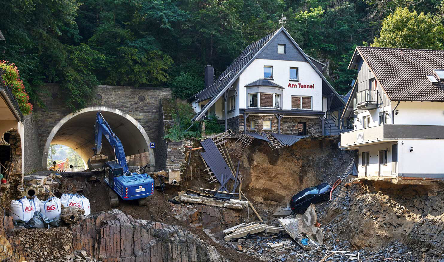Eine Baustelle zeigt einen Erdaushub vor einem Tunnel mit einem Bagger und besch digten Geb uden am Hang. Beschreibung von KI generiert